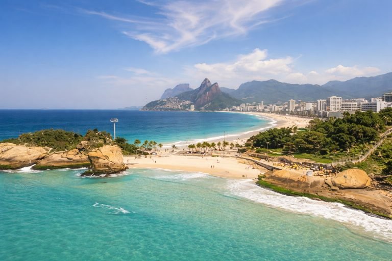 Sunny aerial view of Ipanema Beach in Rio de Janeiro with turquoise water, golden sand, and the Dois Irmãos mountains under bright blue skies. Caption Description