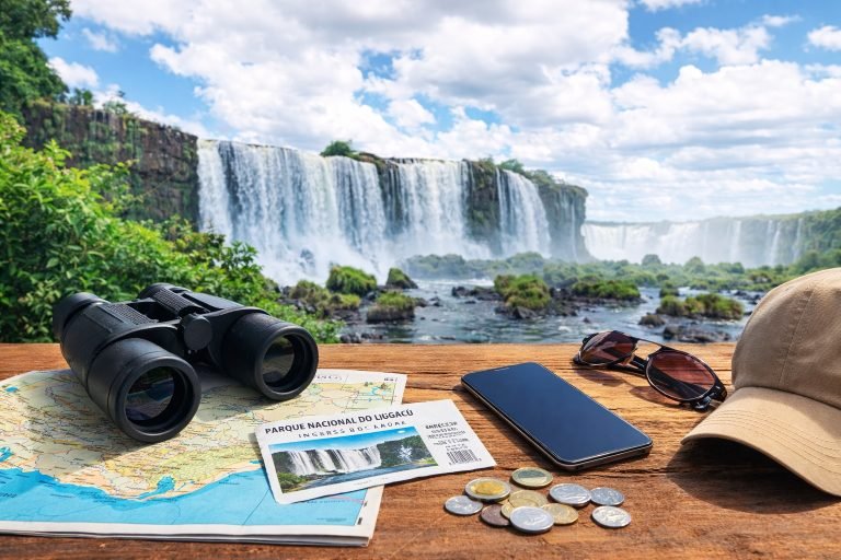 Travel planning essentials including binoculars, map, entrance ticket and Brazilian money on a table with Iguazu Falls in the background