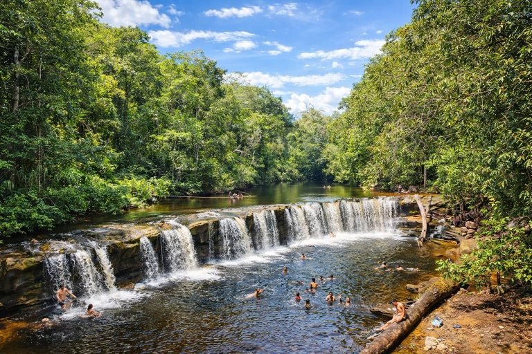 Best Time to Visit Brazil - Sunny Amazon river waterfall with swimmers enjoying clear water surrounded by lush green rainforest under bright blue skies.