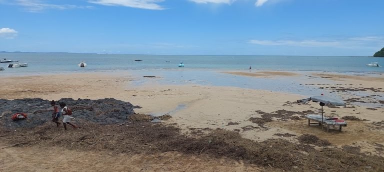 Is Madagascar safe to visit beach scene in Madagascar with local fishermen walking along the shoreline and boats in the ocean