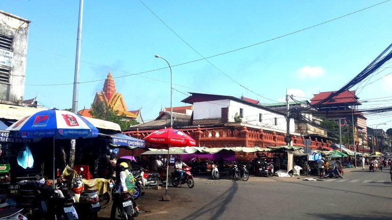 Things to do in Cambodia: busy street scene in Phnom Penh with local market stalls, motorbikes, and temple in the background 📝 Caption 📖 Description