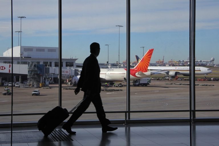 Traveler walking through Sydney Airport terminal with suitcase and airplanes on the runway outside Caption: Description: