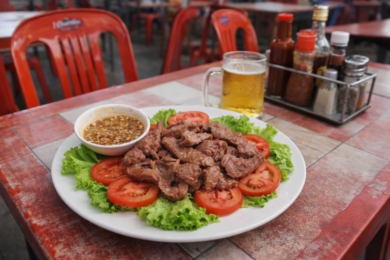 Cambodian food beef lok lak served on a street food table with red plastic chairs and pepper lime dipping sauce Caption Description