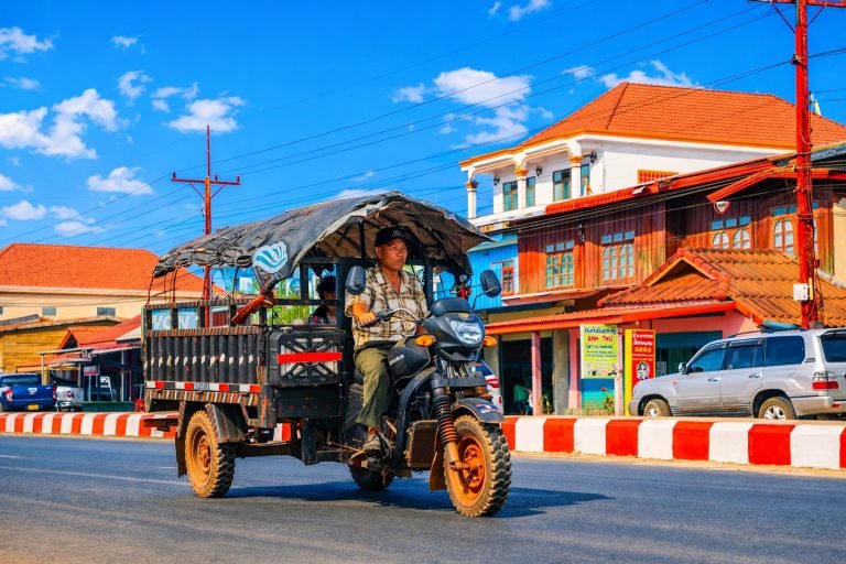 Tuk tuk driving through a sunny street in Battambang Cambodia with colorful buildings and blue skies