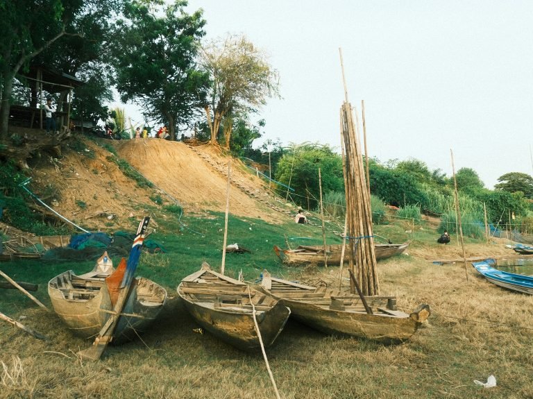 Traditional wooden fishing boats along a riverbank in Battambang Cambodia with rural village surroundings Caption: Description: