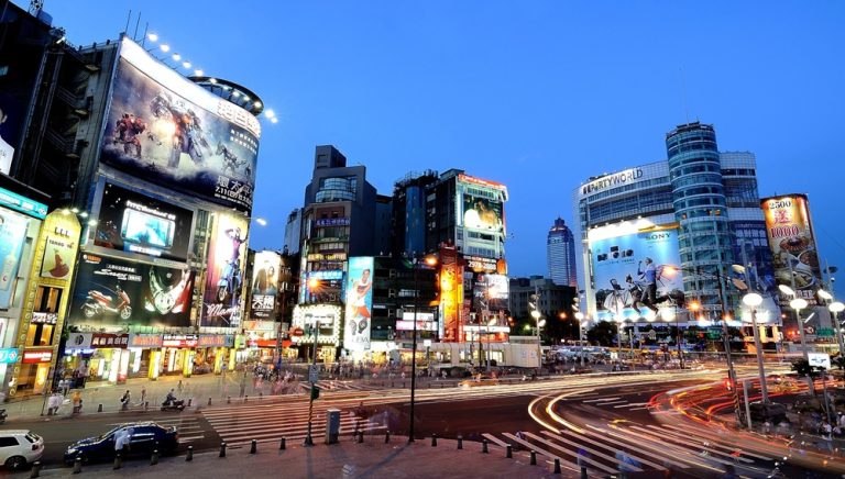 Busy Taipei intersection with neon lights, traffic trails, and illuminated buildings at dusk Caption: Description: