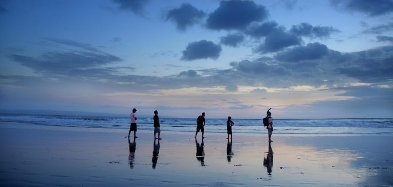Travelers walking along a quiet beach at dusk with ocean reflections and dramatic clouds in Bali Caption: Description: