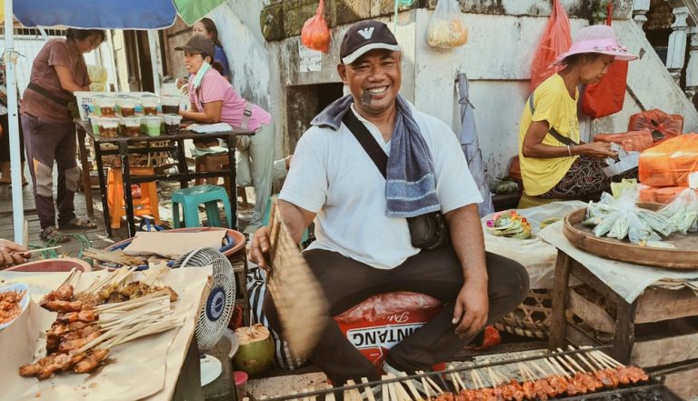 Food in Bali at a traditional street market featuring a local vendor grilling satay skewers over charcoal Caption Description