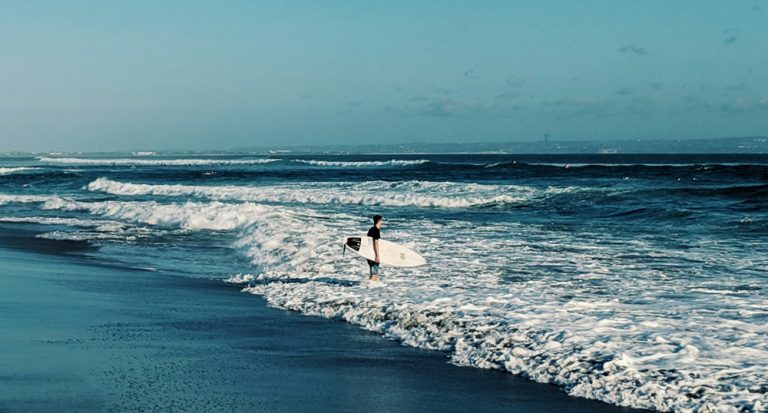 Surfer carrying a surfboard along the shoreline at Batu Bolong Beach in Bali