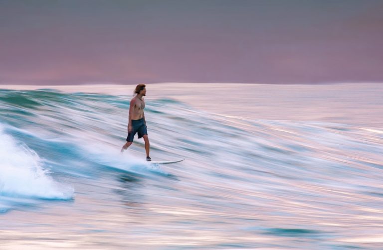 Longboard surfer riding a gentle wave at sunset in Bali, featured in this Bali surf guide. Caption Description