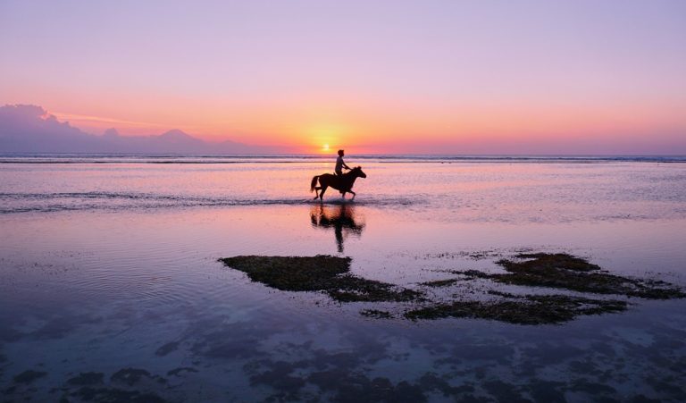Spectacular sunset over Gili Trawangan featured in this Gili Islands travel guide, with a horse rider crossing the shallow shoreline and Mount Agung visible in the distance.