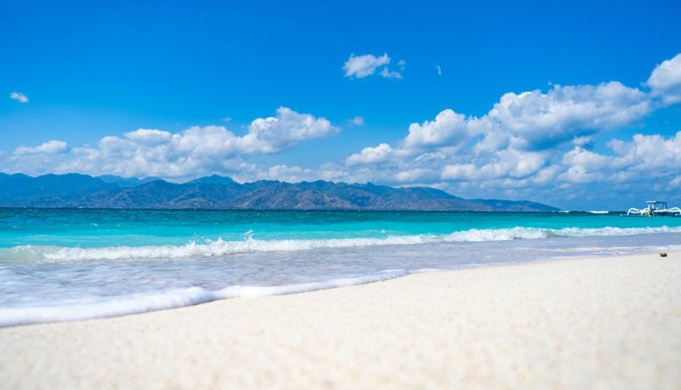 Pristine white-sand beach featured in this Gili Islands travel guide, with turquoise water, gentle waves and views of Lombok in the distance. Caption Description