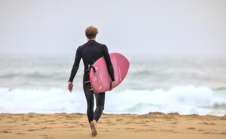 Surfer carrying a pink surfboard along the beach in Bali, featured in this Bali surf guide. Caption Description