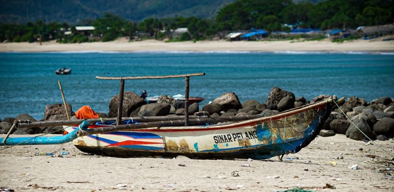 Traditional Indonesian fishing boat resting on a tropical beach with turquoise water and a sandy coastline in Indonesia. Caption Description