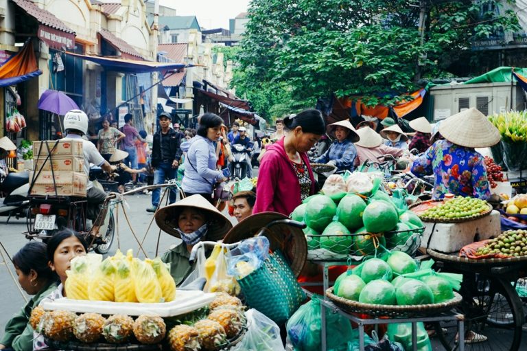 Busy local street market with fruit vendors showing everyday food prices for a Vietnam travel budget Caption: Fruit vendors and shoppers fill a busy traditional street market in Vietnam. Busy local street market with fruit vendors showing everyday food prices for a Vietnam travel budget