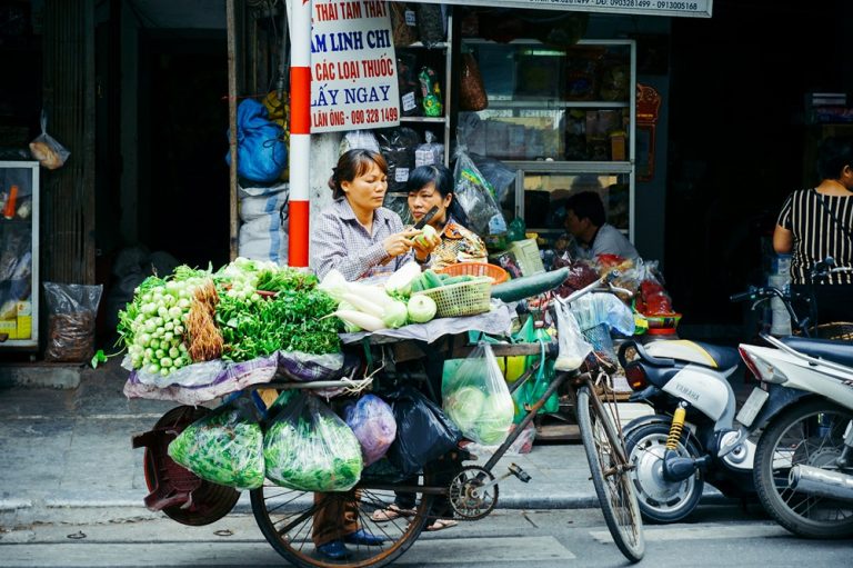 Woman preparing fresh vegetables beside a bicycle loaded with produce on a busy Vietnamese street.