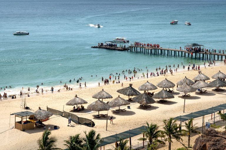 Visitors swimming at a sandy beach in Nha Trang with thatched umbrellas, a boat pier and turquoise sea