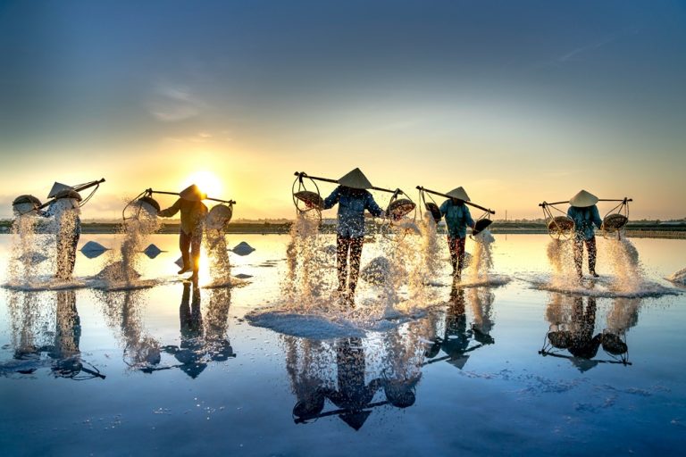 Salt workers carrying baskets across shallow salt fields near Nha Trang, Vietnam, at sunrise